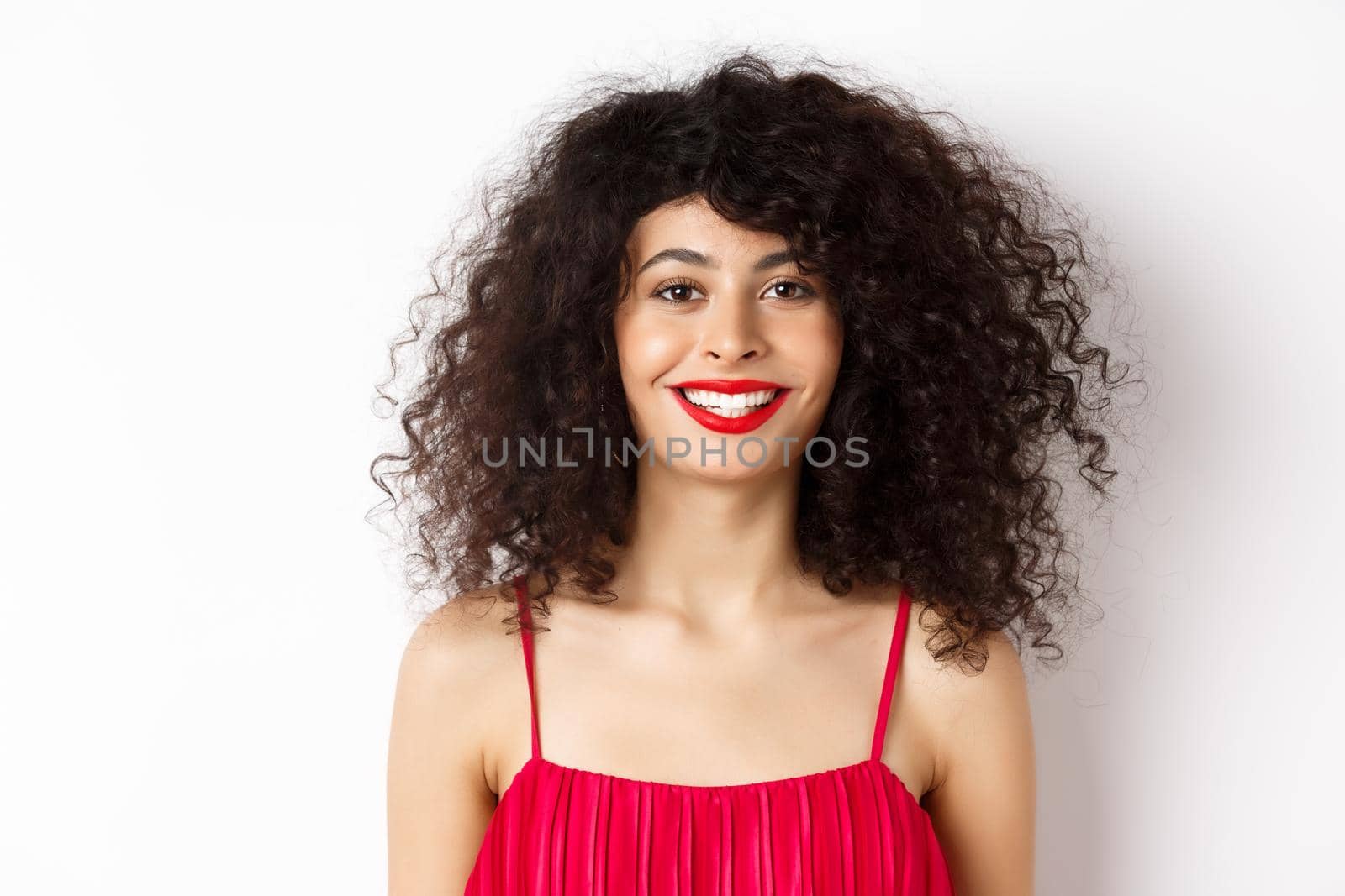 Close-up of beautiful woman with curly hairstyle, wearing red dress and lipstick, smiling happy at camera, white background.