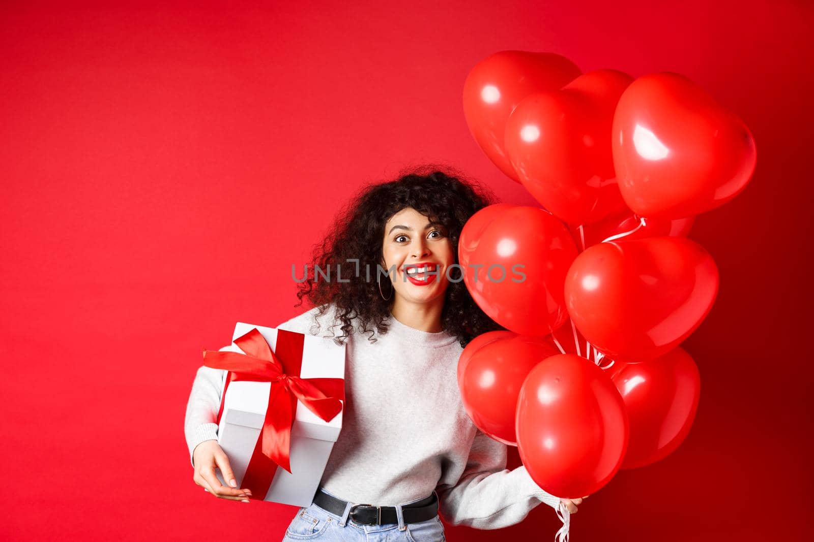 Holidays and celebration. Happy birthday girl holding gift and posing near party helium balloons, smiling excited at camera, red background by Benzoix