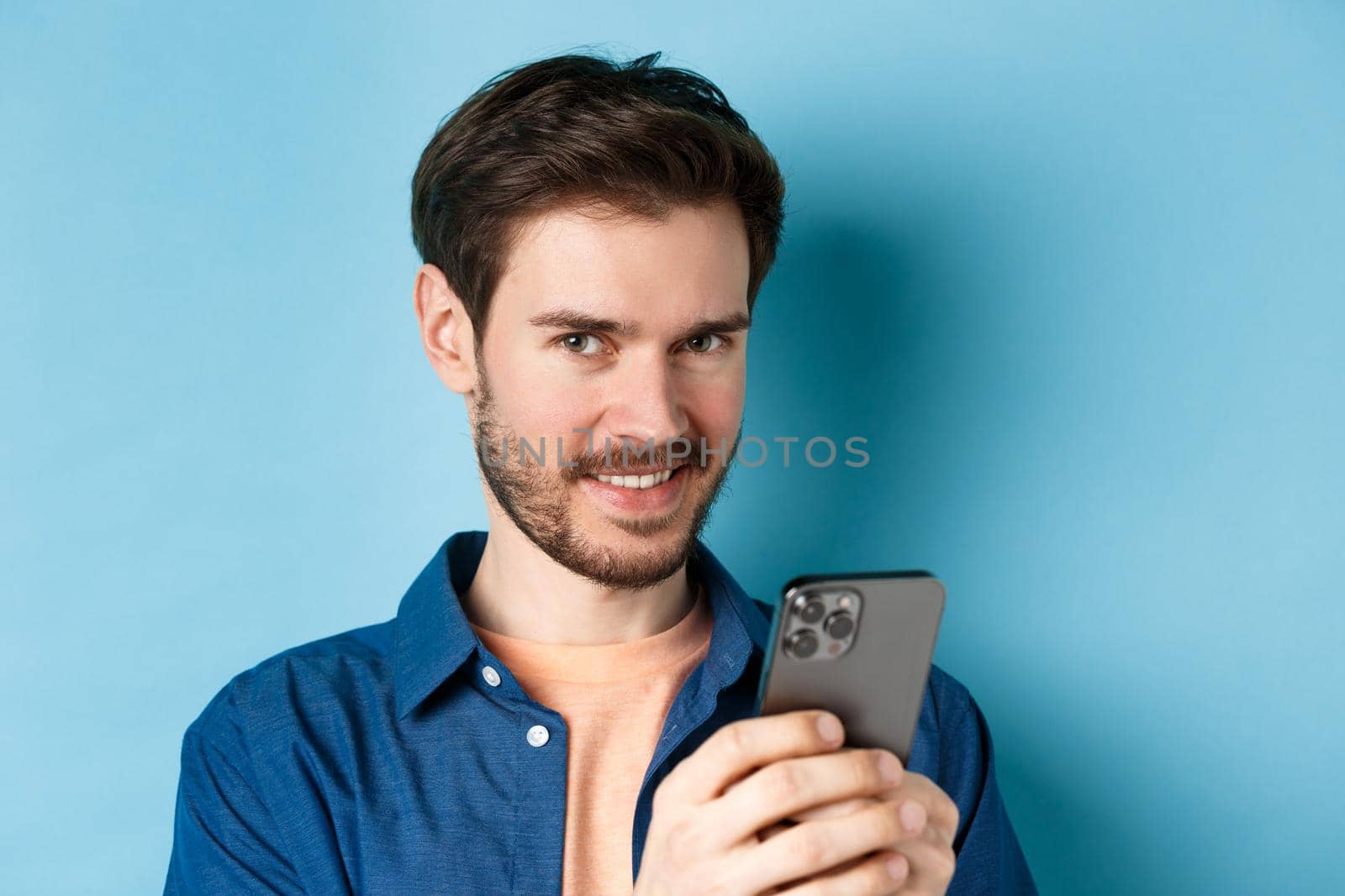 Close-up of attractive caucasian man holding mobile phone and smiling at camera, standing in casual outfit on blue background by Benzoix