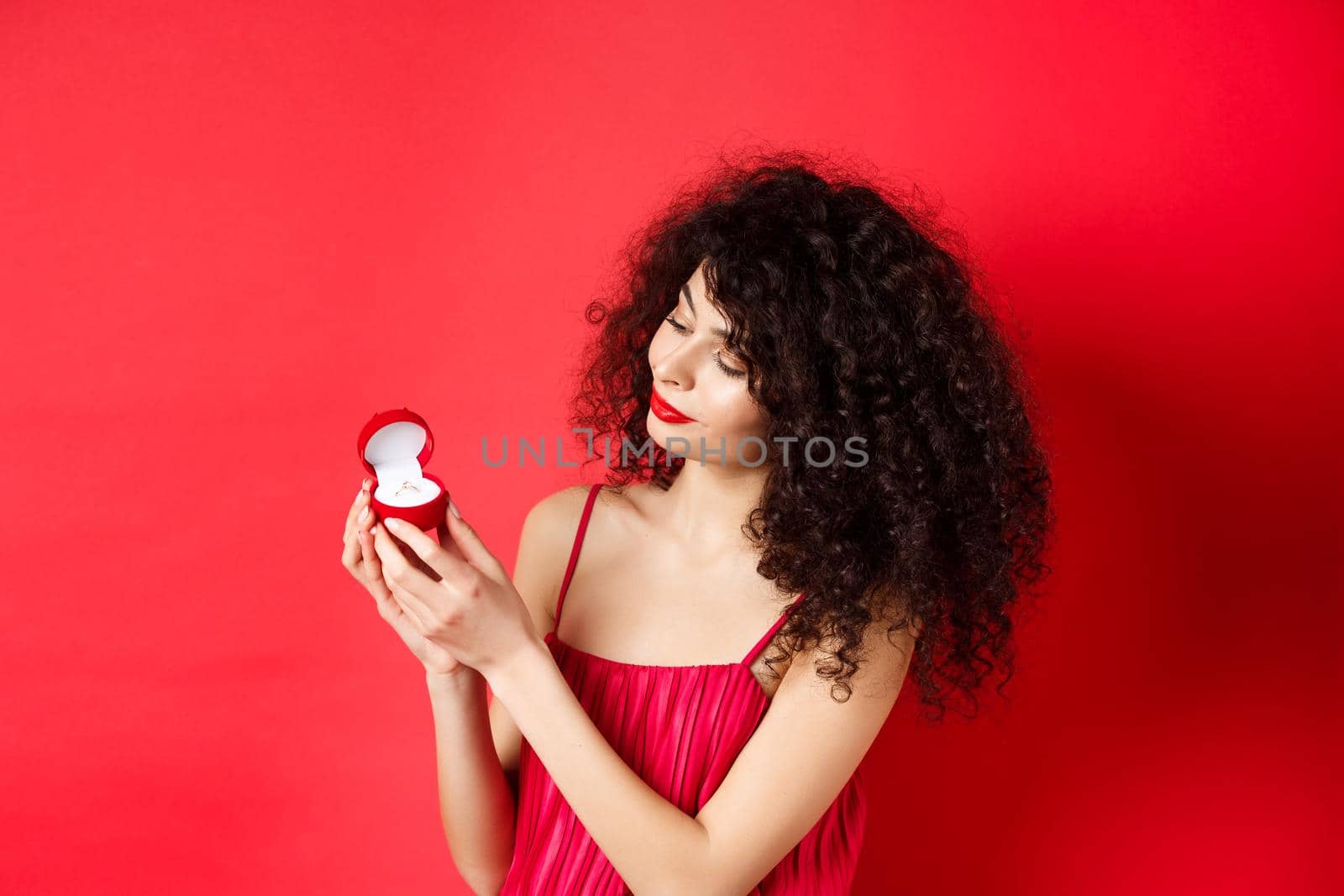 Romantic curly-haired woman in red dress, looking happy at engagement ring, become a bride, standing on studio background. Copy space