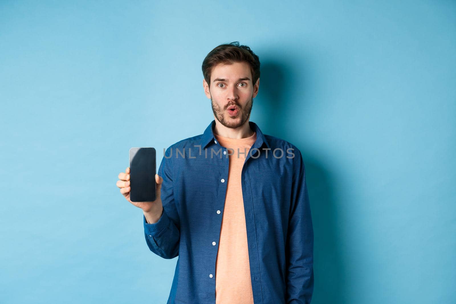 Impressed caucasian male model showing smartphone screen and look amazed, talking about online offer, standing on blue background.
