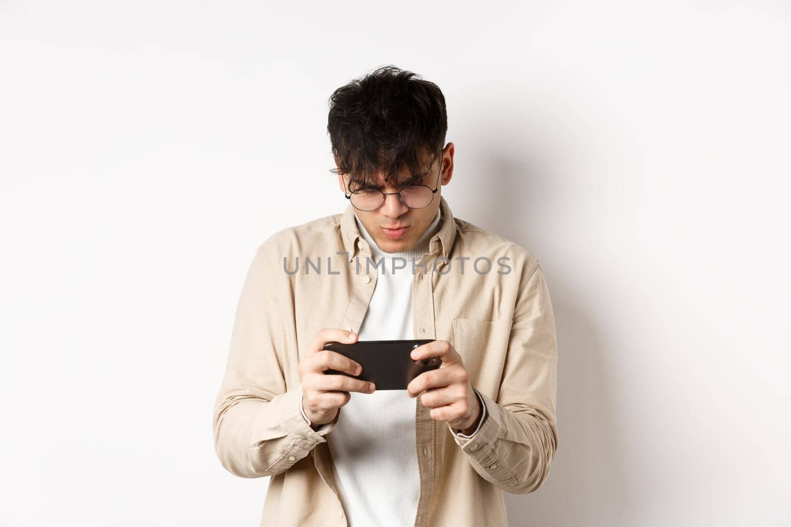 Handsome young man stare at mobile screen and playing video game on smartphone, standing on white background by Benzoix