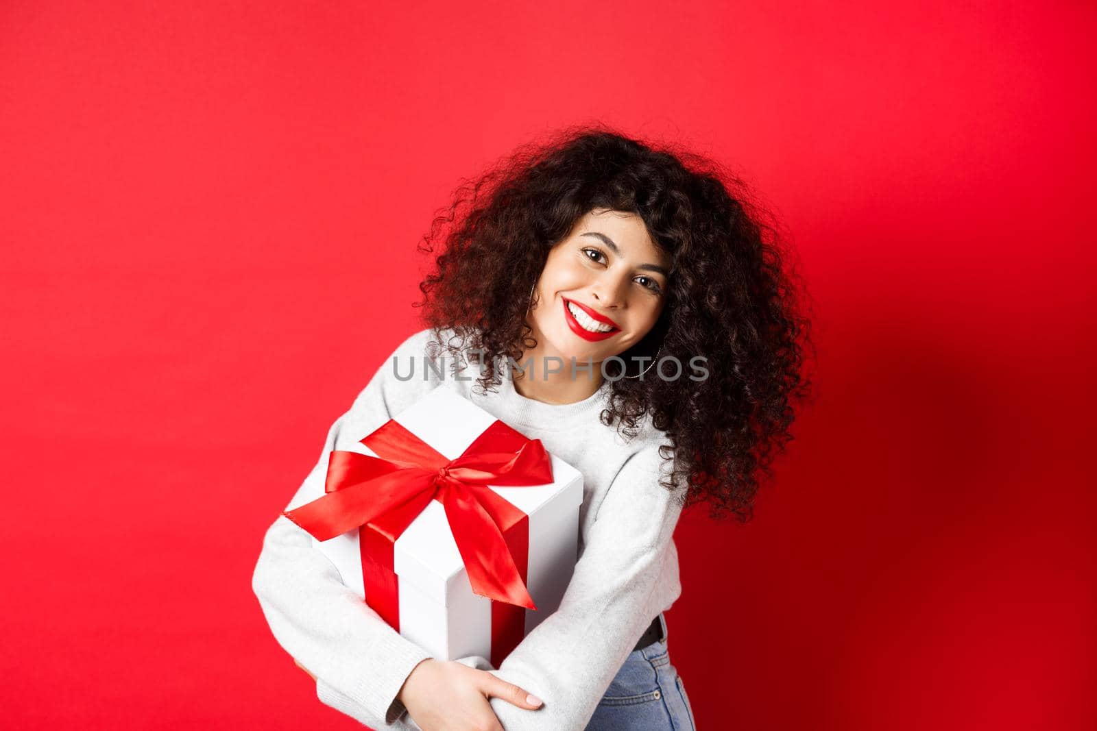 Celebration and holidays concept. Happy woman holding birthday gift and smiling at camera, standing in casual clothes, red background.