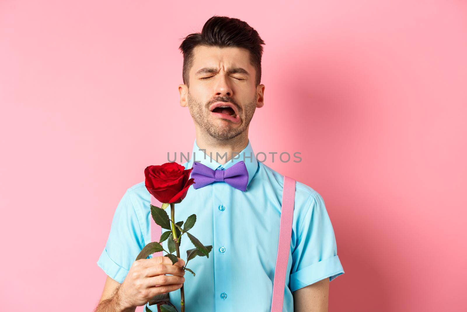 Heartbroken guy in funny bow-tie crying over girlfriend, standing alone with red rose on pink background and sobbing, break-up on Valentines day by Benzoix
