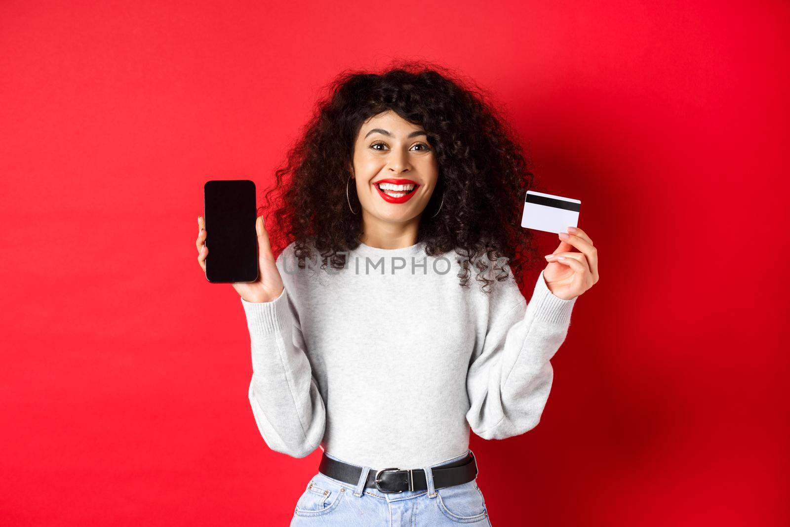 E-commerce and online shopping concept. Cheerful woman smiling, showing plastic credit card and empty smartphone screen, standing on red background.