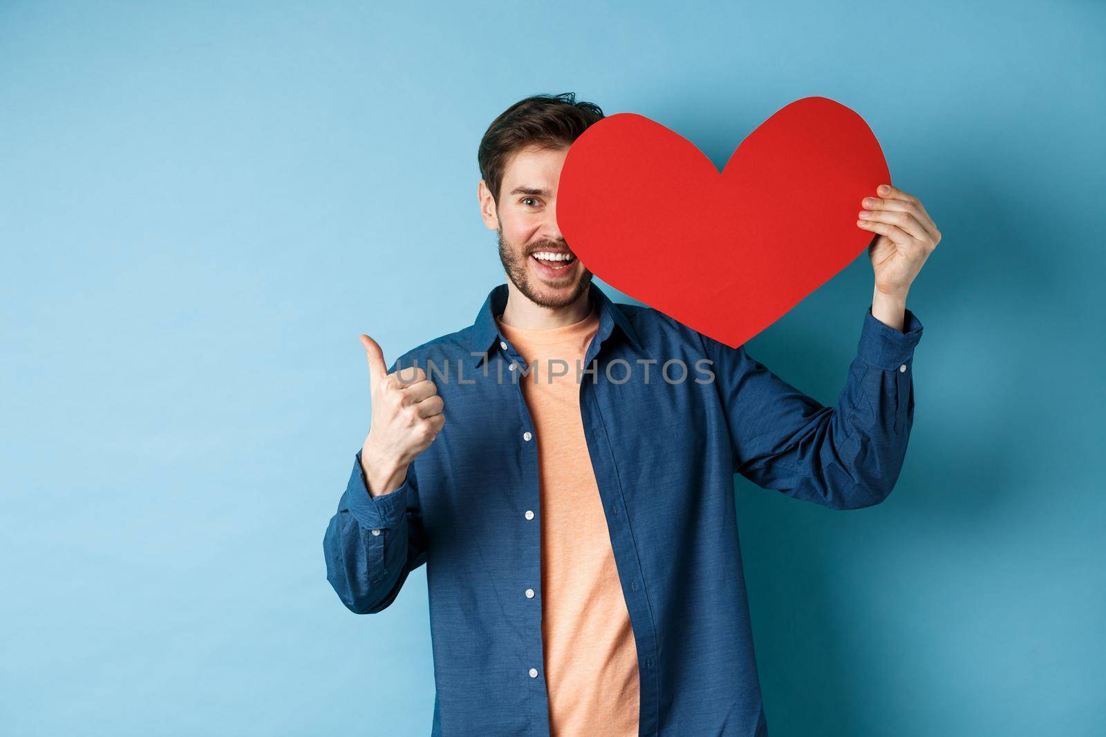 Happy man showing valentines heart and thumbs-up gesture, standing over blue background.