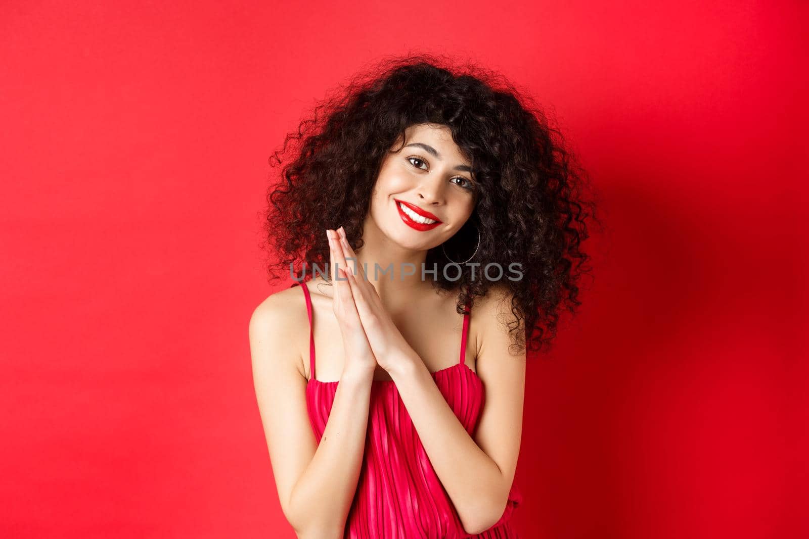 Tender woman with curly hair, wearing red dress, looking at something lovely, thanking you, standing over studio background. Copy space
