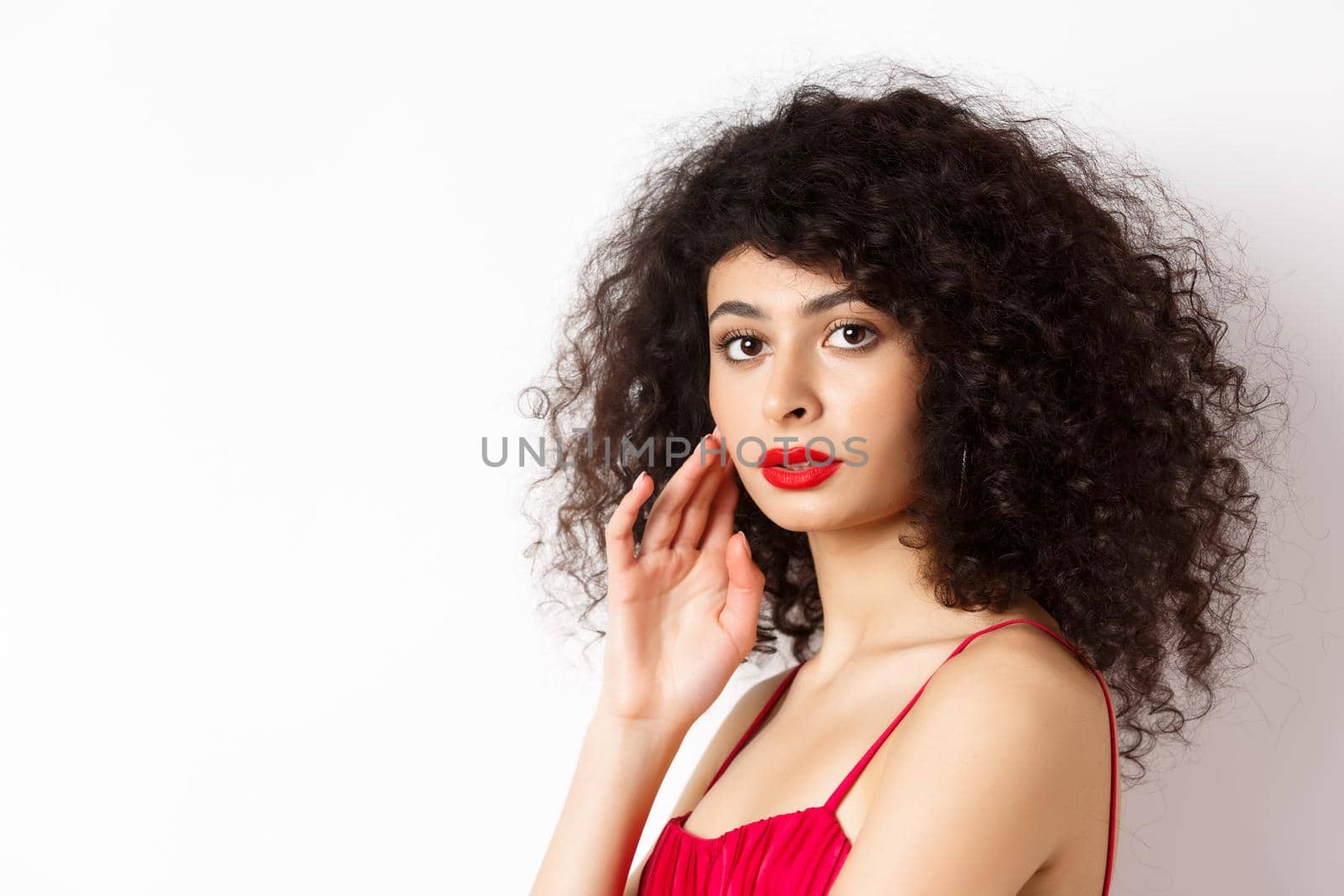 Close up of elegant woman with curly hair and red lips, looking at camera and touching face, standing in red dress on white background by Benzoix