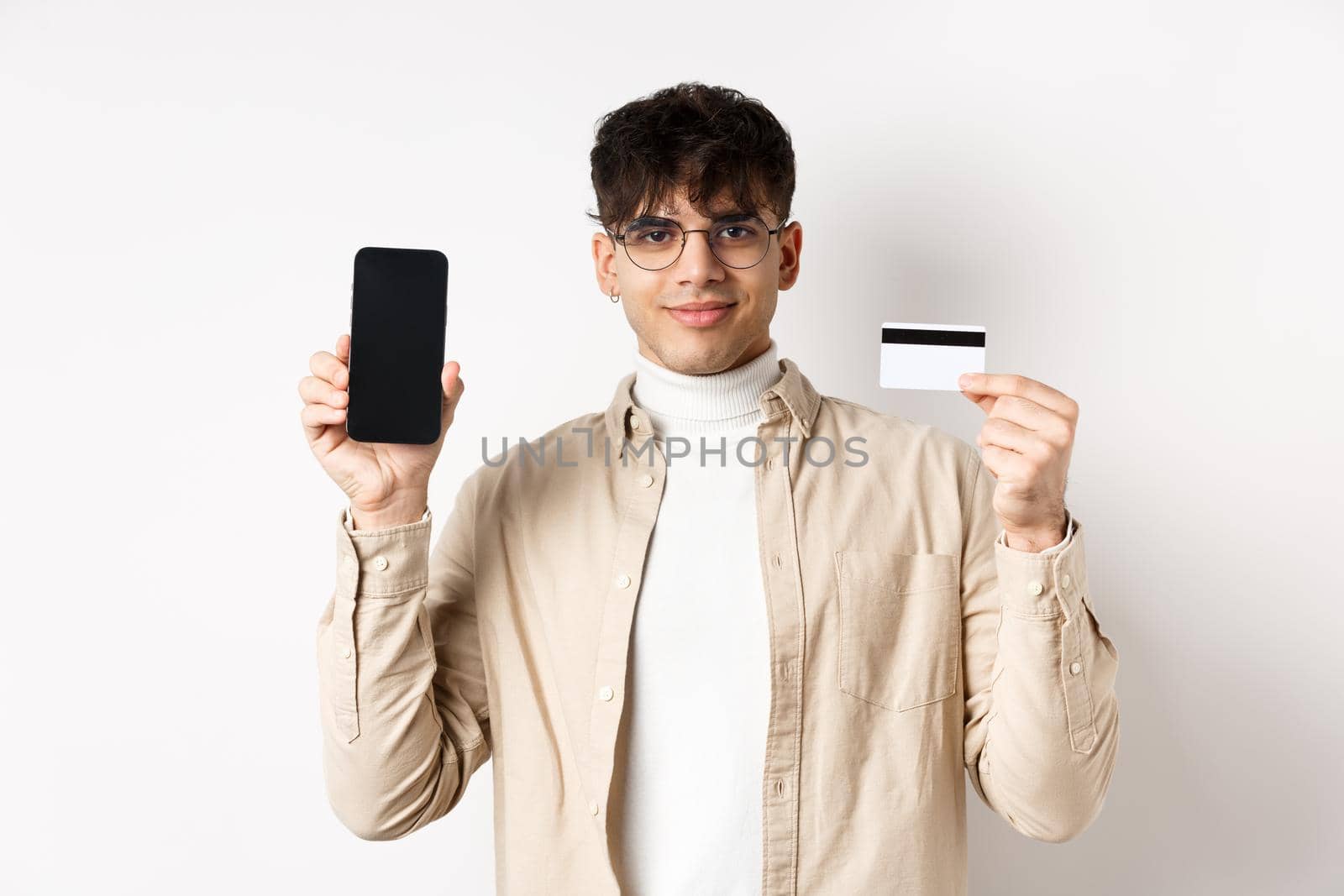 Online shopping. Young modern guy showing plastic credit card and empty smartphone screen, demonstrate account, standing on white background.