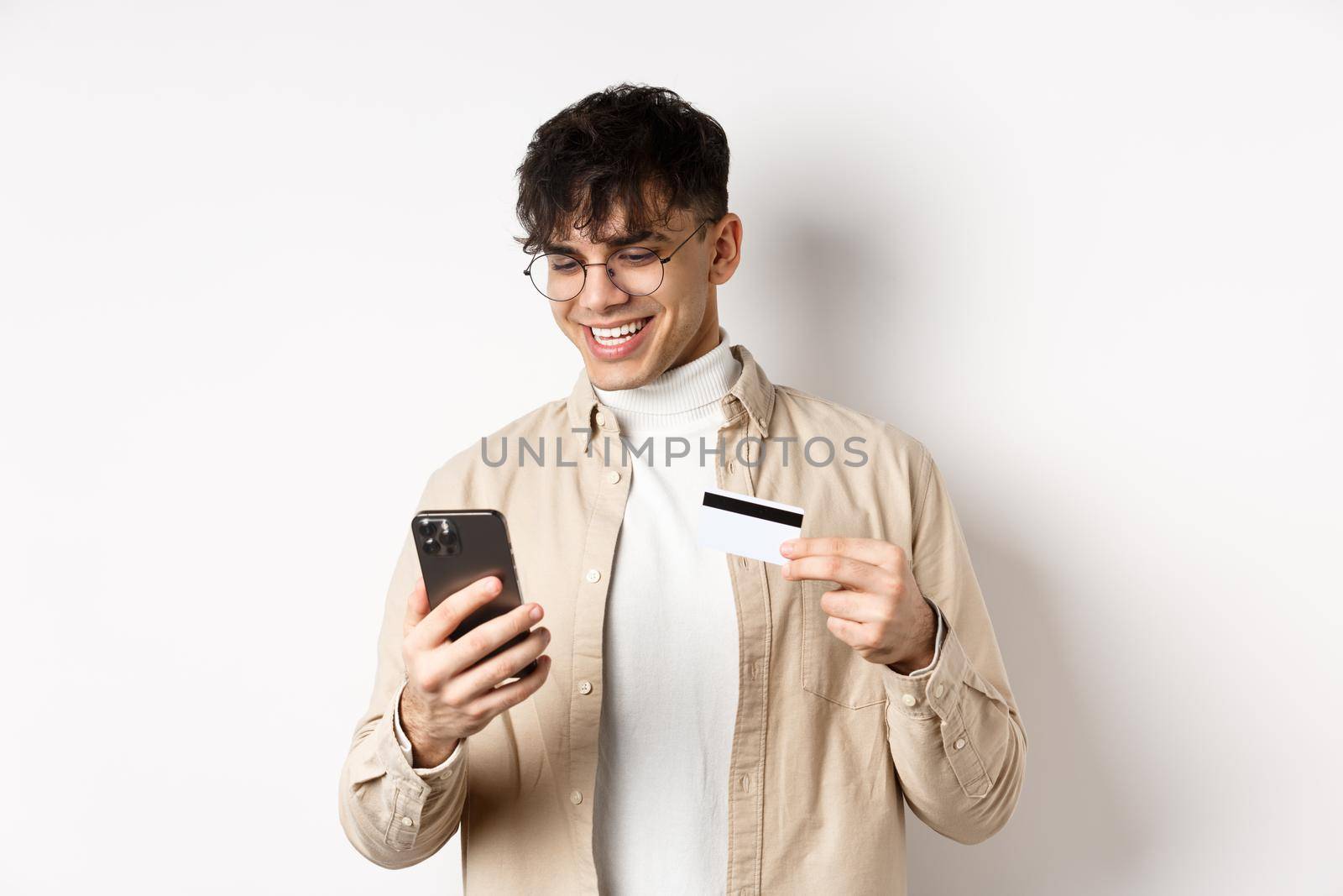 Online shopping. Happy young man using smartphone and plastic credit card, paying in internet, standing on white background.