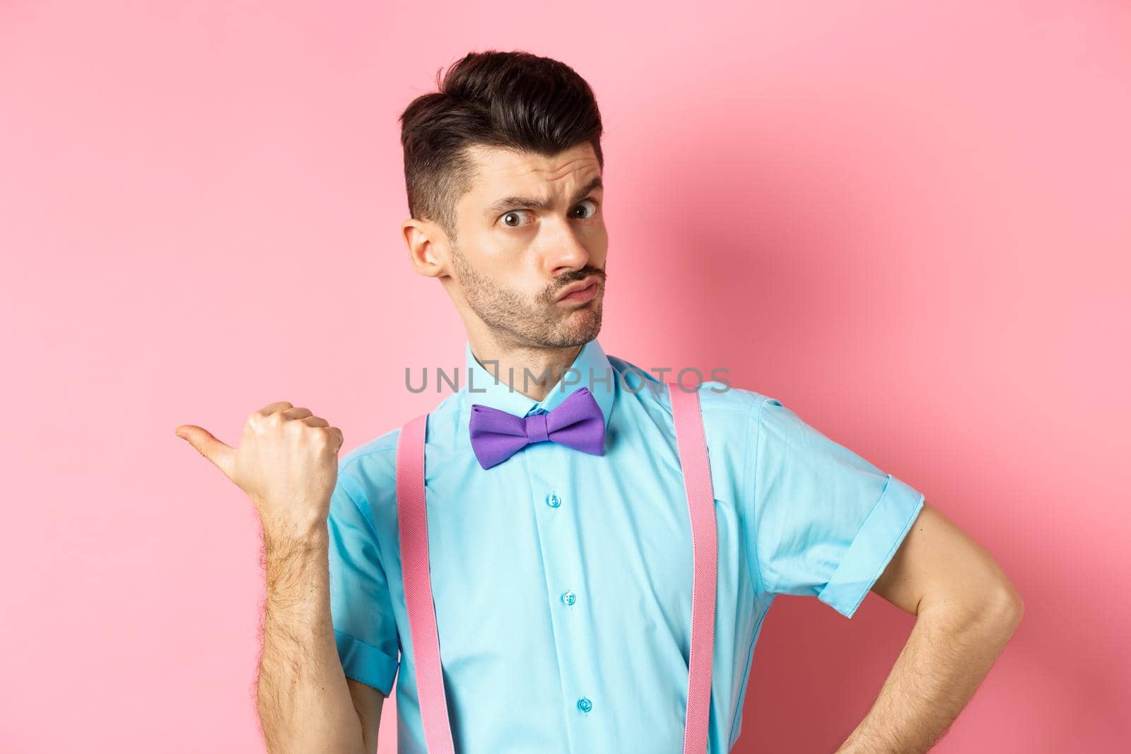 Serious-looking guy in suspenders suggessting to go outside, pointing left and looking confident at camera, starting fight, standing over pink background by Benzoix