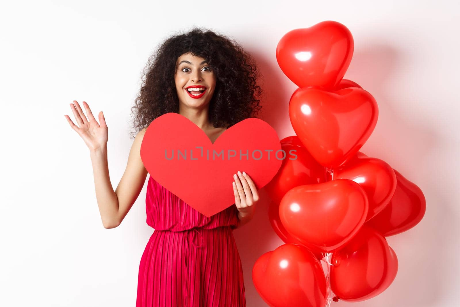 Valentines day and love concept. Cheerful young woman in elegant red dress, standing near romantic balloons and holding big red heart cutout, waving hand to say hi, waiting for date.