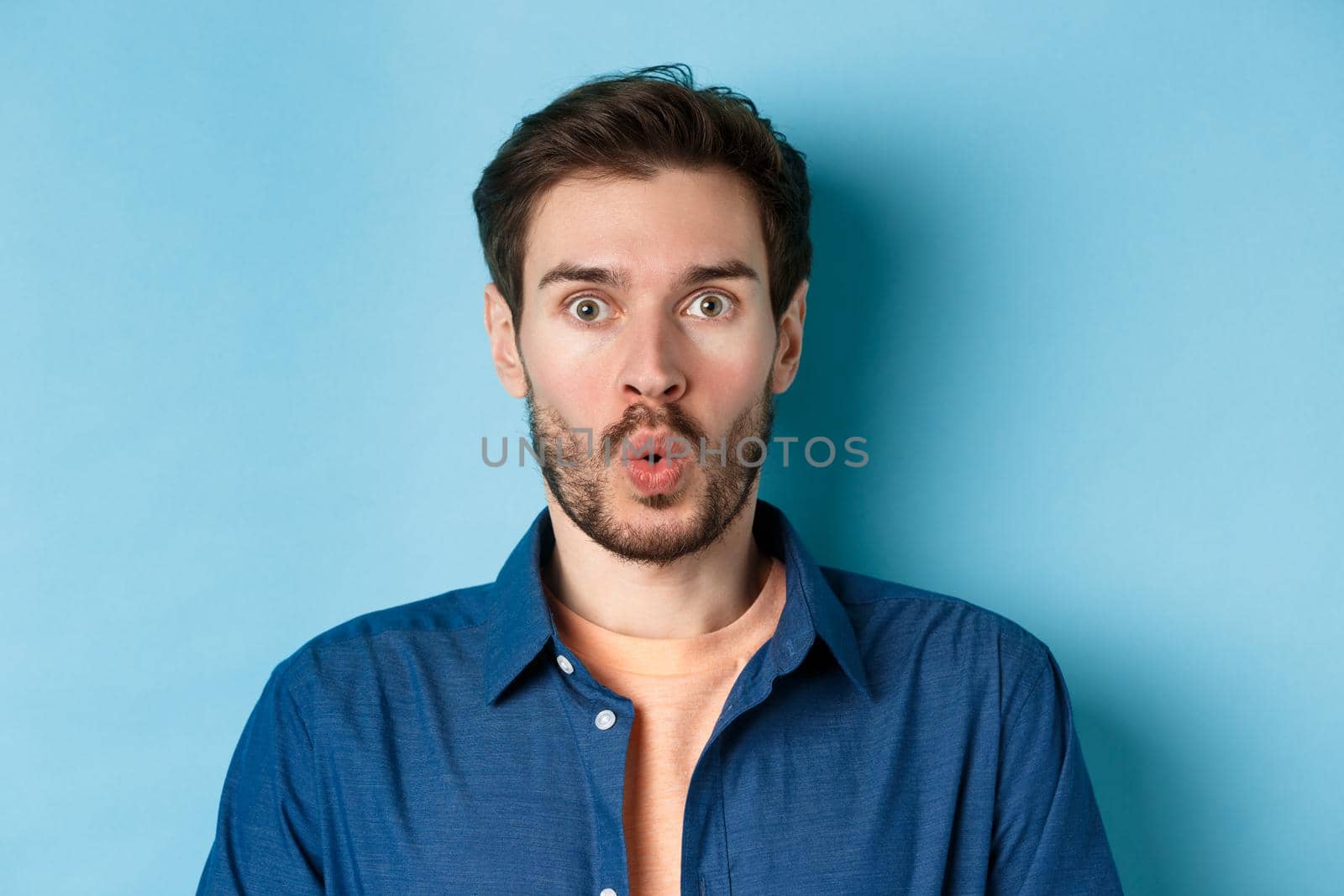 Close up portrait of amazed and surprised man saying wow, staring impressed at camera, checking out cool offer, standing on blue background.