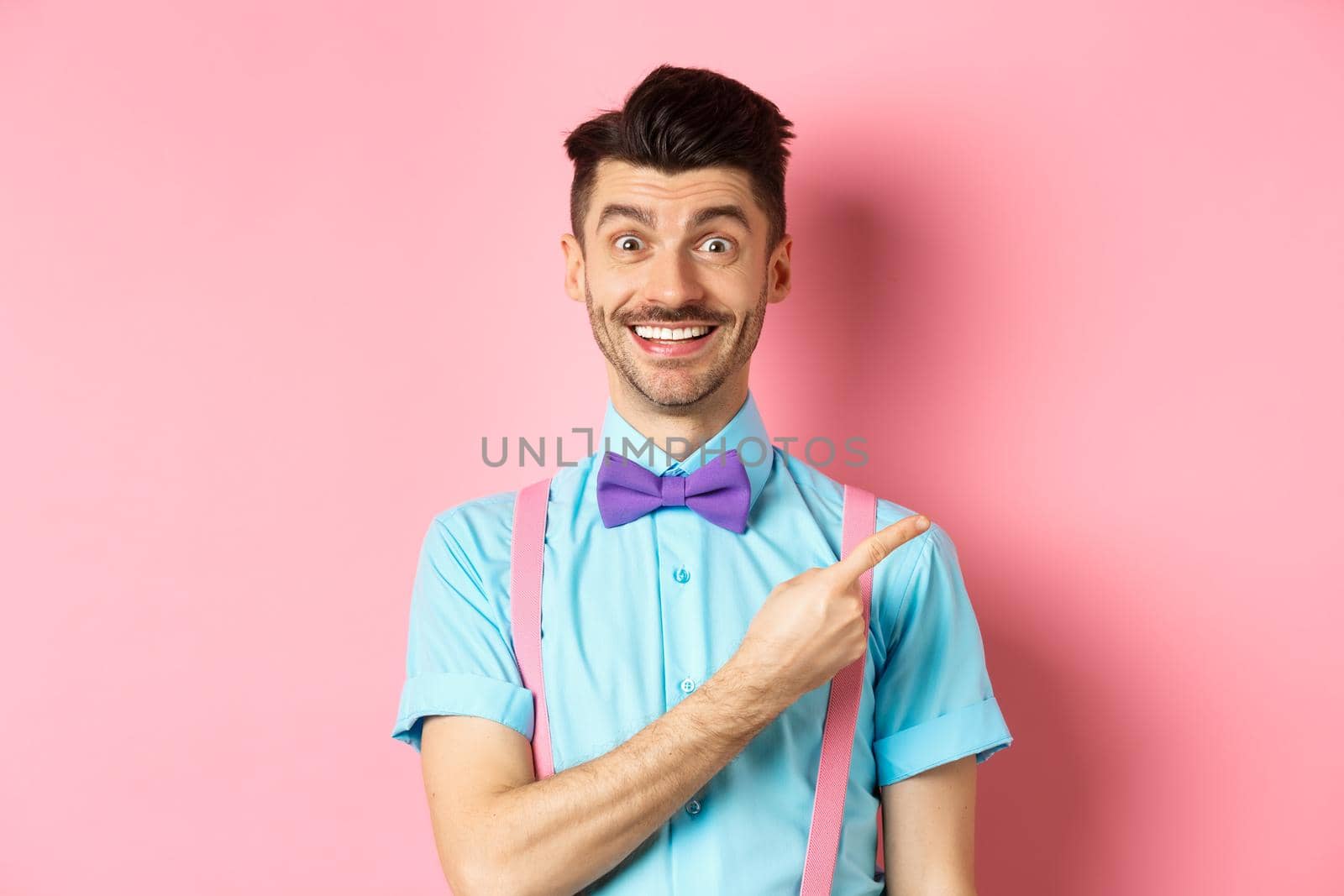 Handsome caucasian guy with bristle and moustache pointing finger right aside, showing advertisement, standing over pink background in classy bow-tie by Benzoix