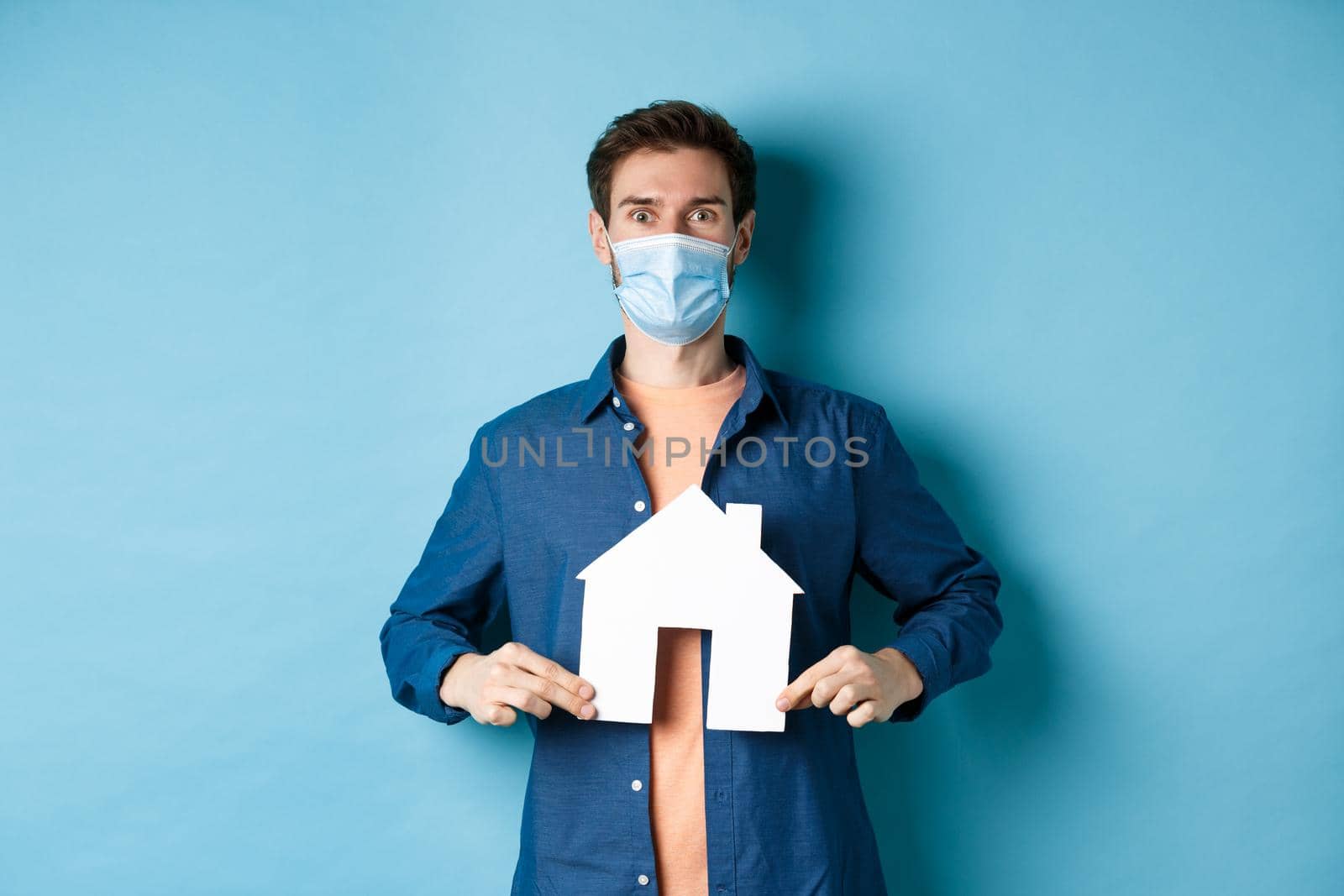 Real estate and covid concept. Happy young man in face mask demonstrate paper house cutout, looking surprised at camera, blue background.