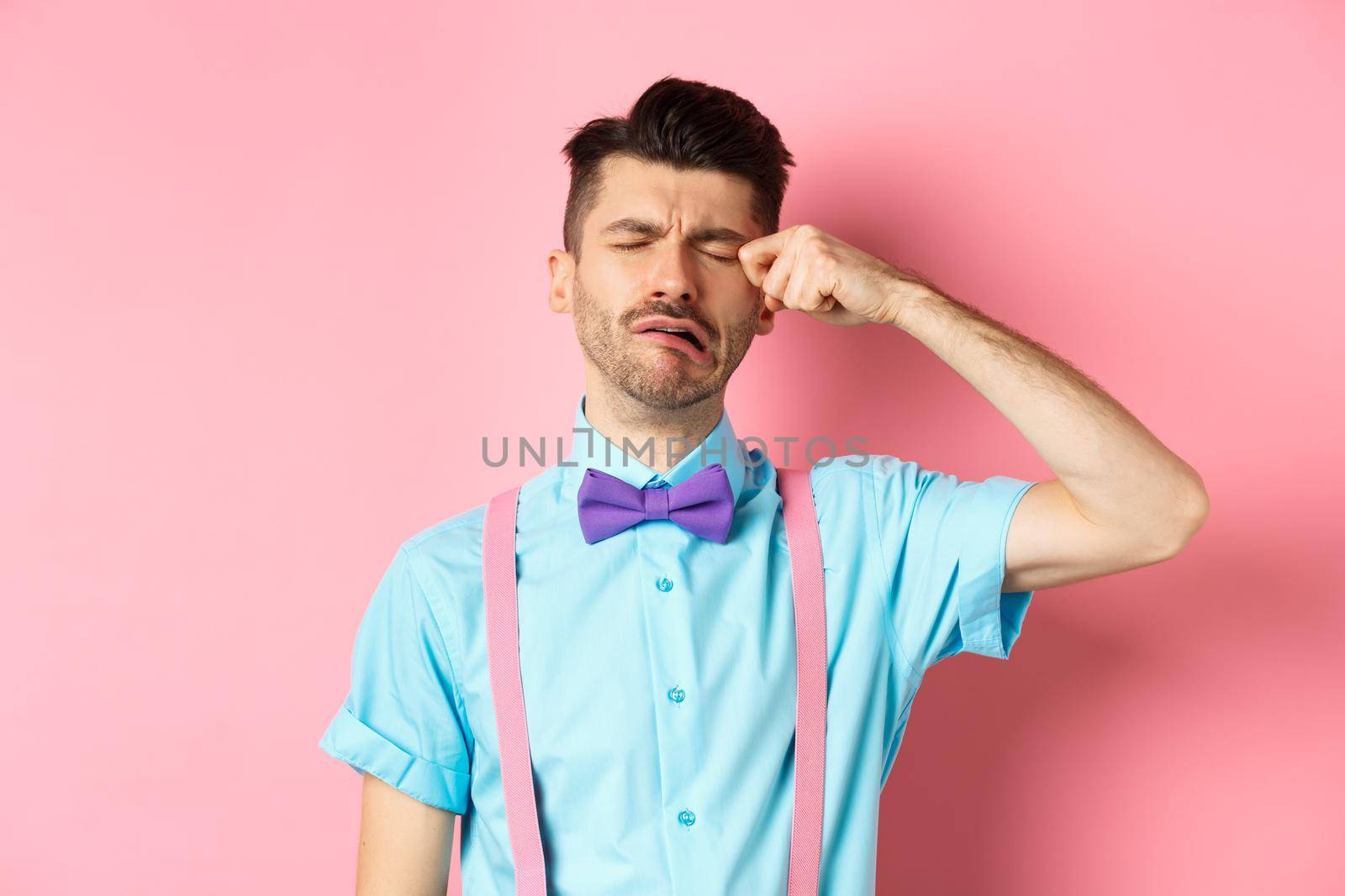 Image of heartbroken guy crying and wiping tear off face, sobbing and feeling sad or lonely, standing upset on pink background.