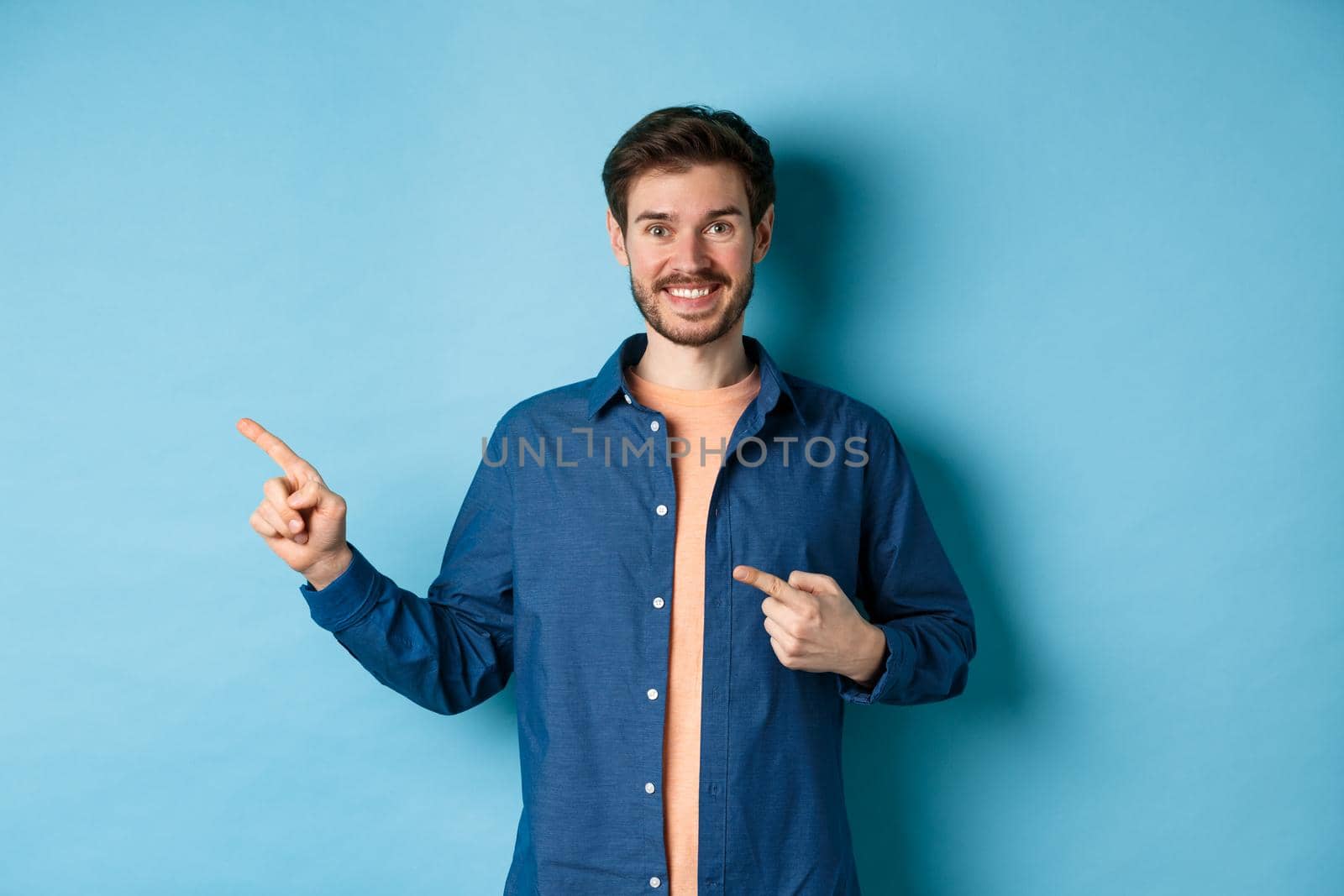 Happy smiling man pointing fingers left at logo, showing advertisement, standing on blue background by Benzoix