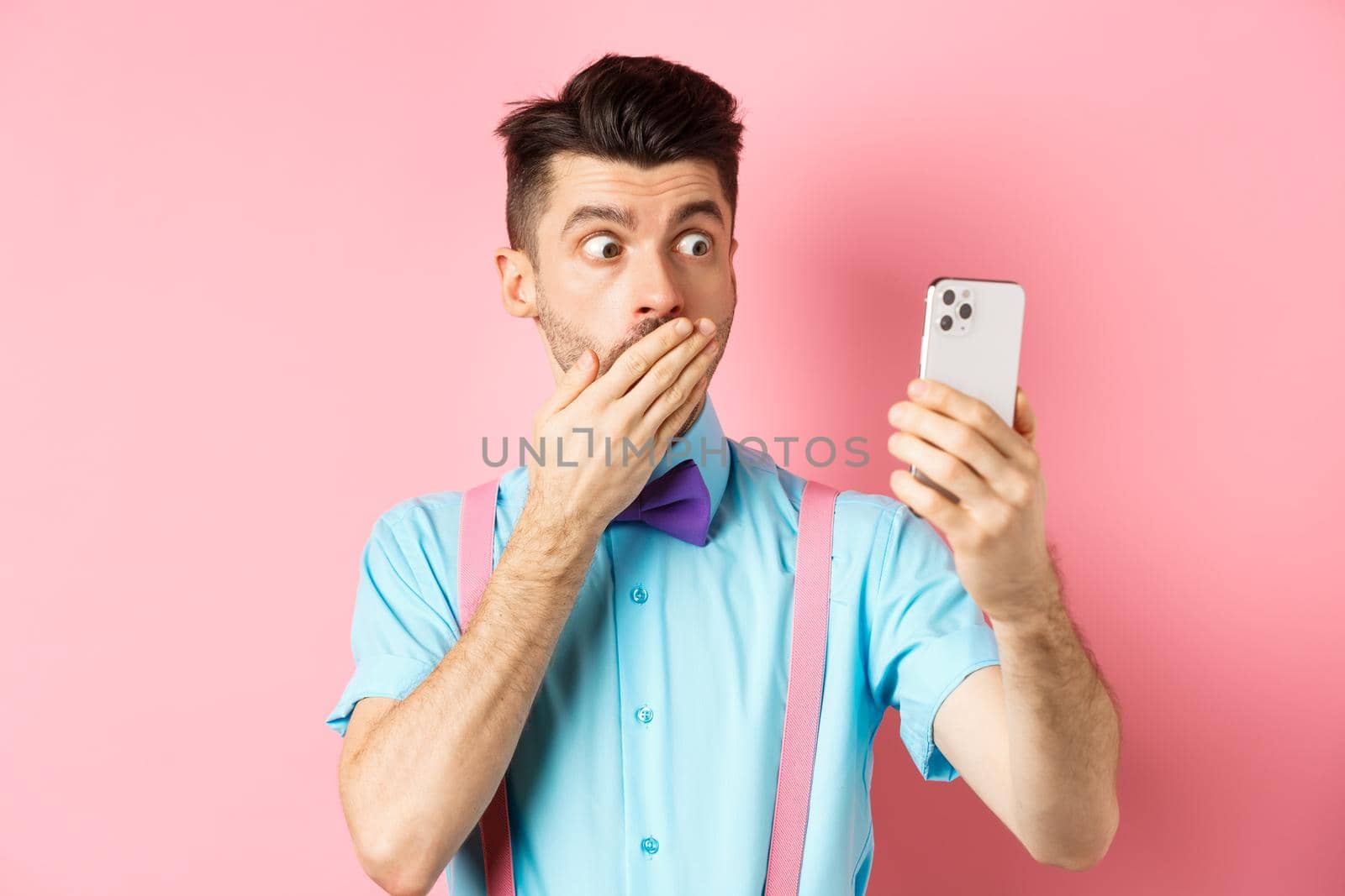 Technology concept. Silly young man looking shocked at smartphone screen while having video chat, standing on pink background and cover mouth with hand by Benzoix