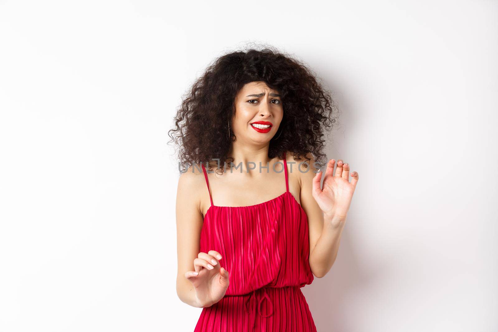 Young woman cringe of something creepy, grimacing and recoil from disgusting awful thing, standing in red dress on white background.