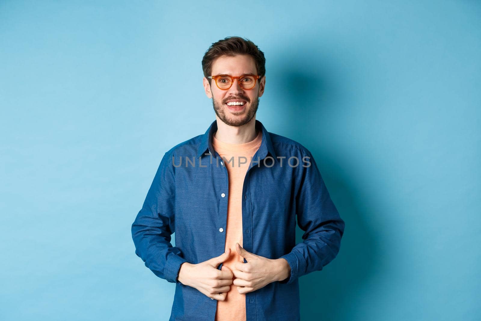 Handsome bearded man smiling pleased, wearing new glasses, standing on blue background.