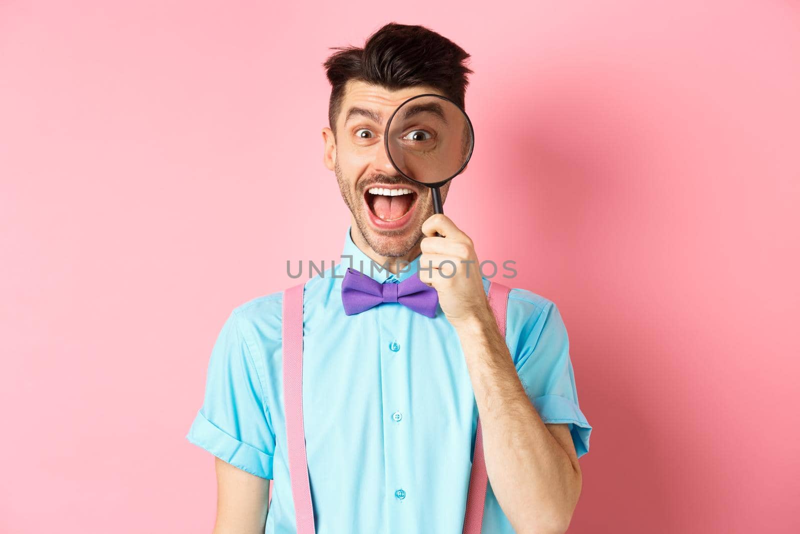 Funny young man in bow-tie look through magnifying glass and showing tongue, smiling happy, having fun on pink background.