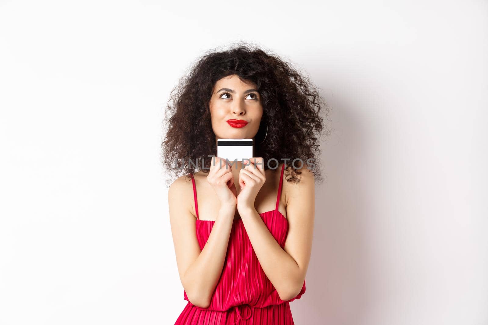 Silly young woman looking romantic, holding plastic credit card and thinking of shopping, standing over white background. Copy space