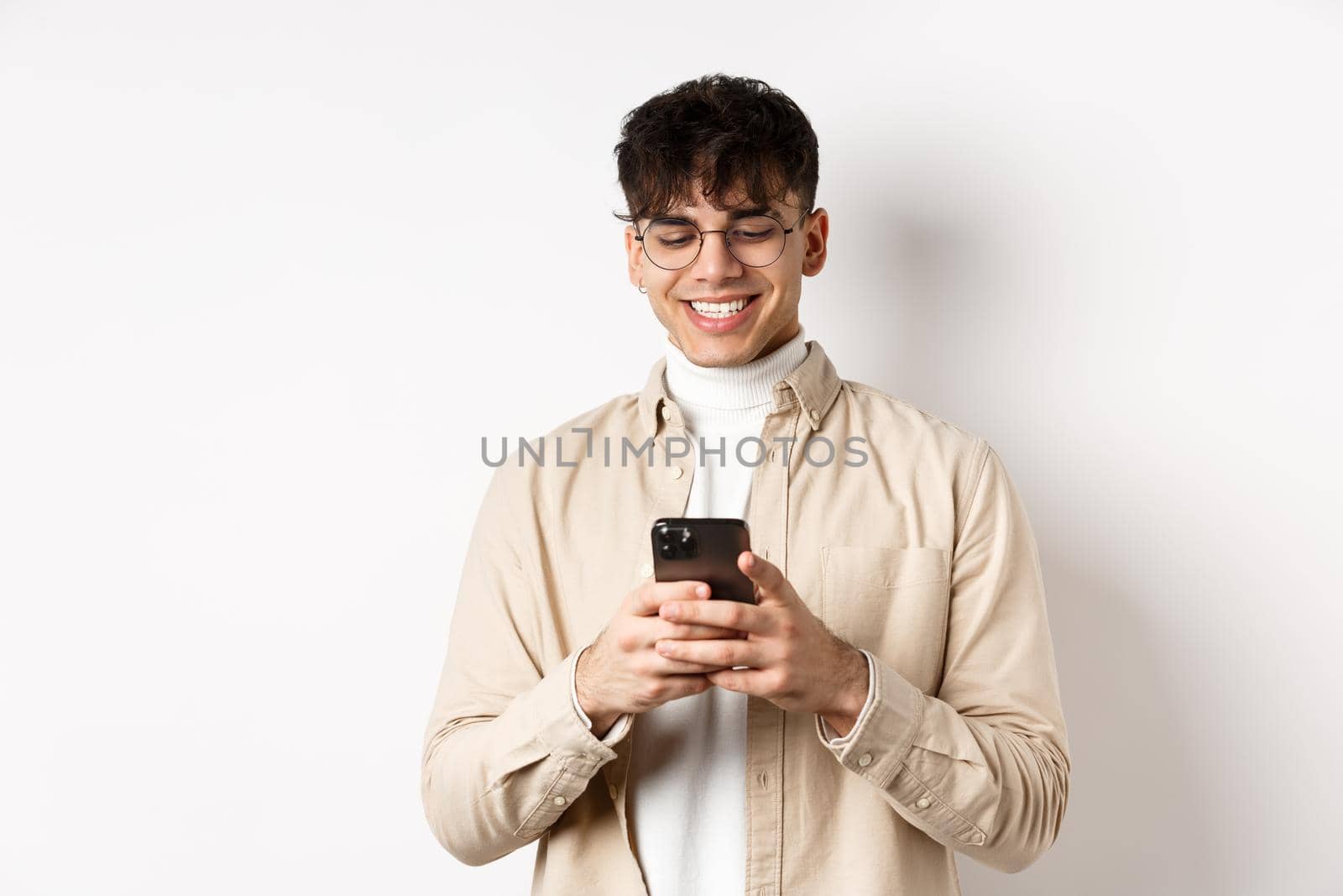 Real people. Natural young man reading message on mobile phone, smiling and looking at smartphone screen, standing on white background.