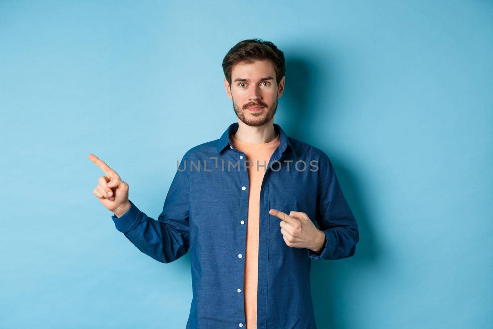 Handsome young man in shirt, pointing fingers left at promo, showing logo, standing on blue background.