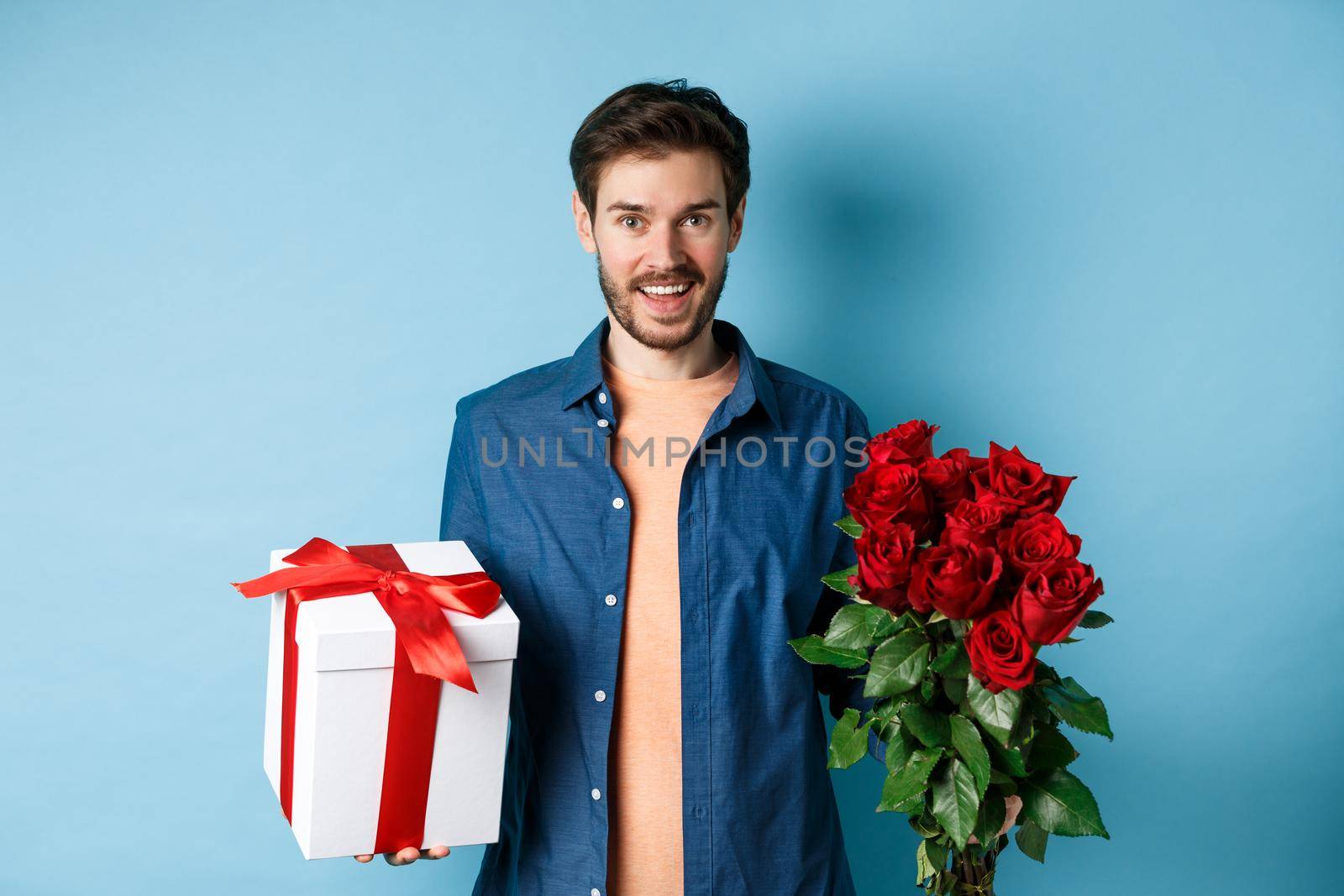 Love and relationship concept. Happy young man bring flowers and gift on romantic date. Boyfriend wish bouquet of roses and present standing on blue background.