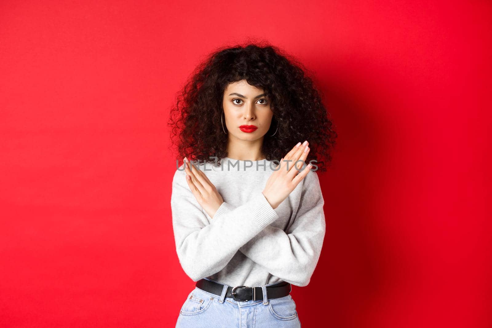 Serious caucasian woman with curly hair say no, making cross gesture to stop or prohibit something bad, standing displeased on red background by Benzoix