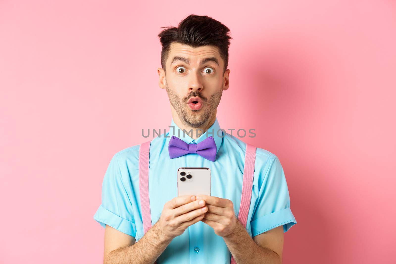 Online shopping. Intrigued guy in bow-tie, checking out internet promo offer on mobile phone, say wow at camera, holding smartphone, standing on pink background by Benzoix