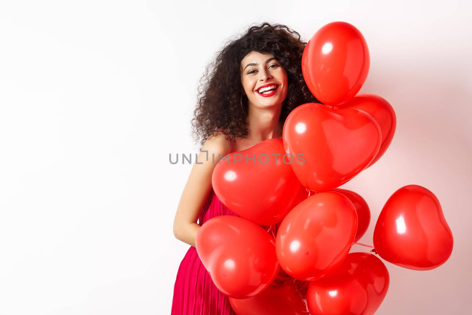 Beautiful caucasian woman with curly hair, wearing date dress, holding romantic red hearts balloons and laughing on Valentines day, standing happy on white background.