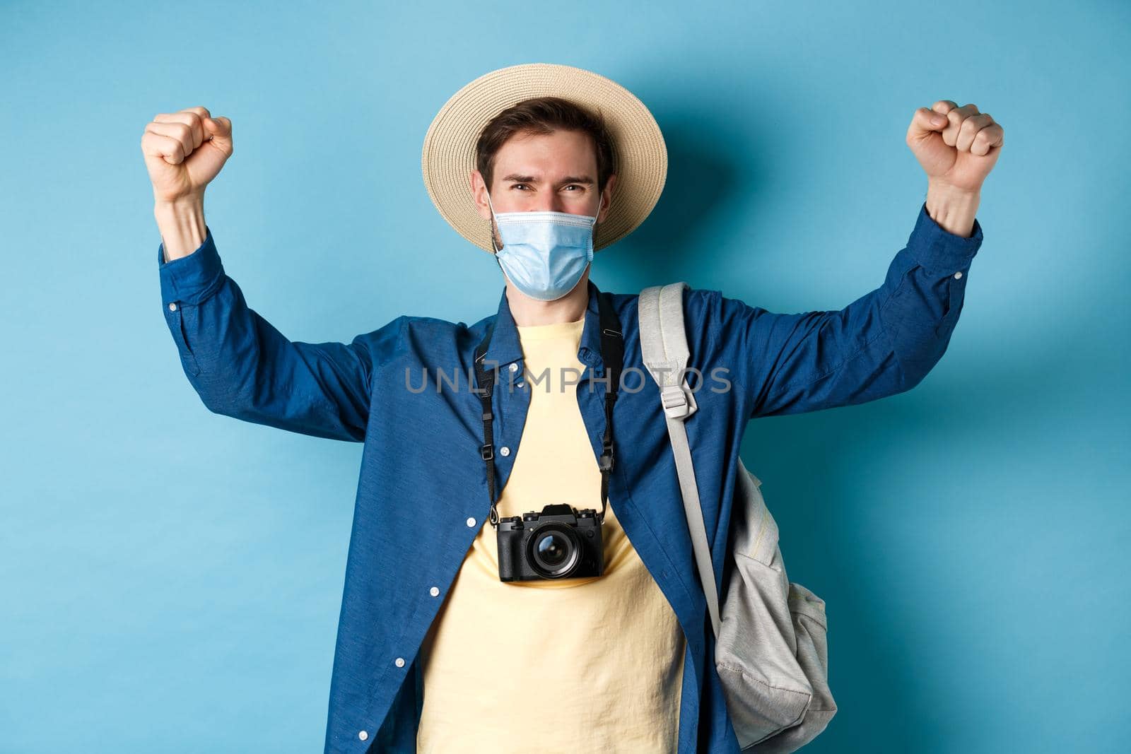 Covid-19 and summer holidays concept. Happy smiling tourist celebrating vacation, wearing face mask, shouting yes with joy and raising hands up, standing on blue background.