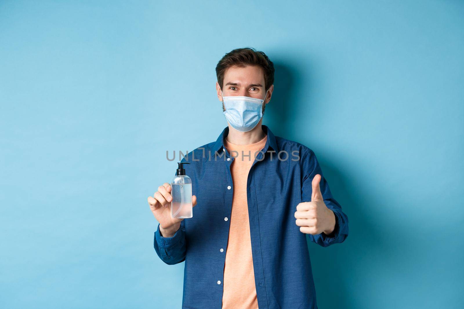 Coronavirus, quarantine and social distancing concept. Happy male model in face mask showing hand sanitizer and thumb up, praise good product, blue background.