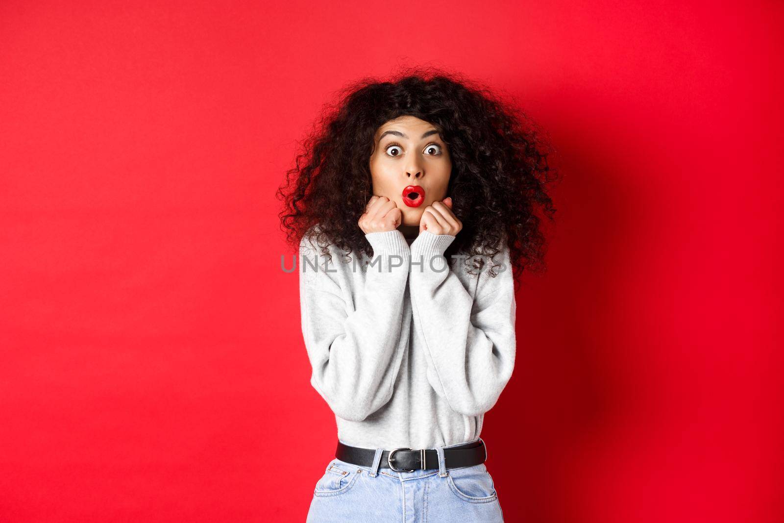 Surprised young woman looking with disbelief and amazement at camera, saying wow and standing in awe on red background by Benzoix