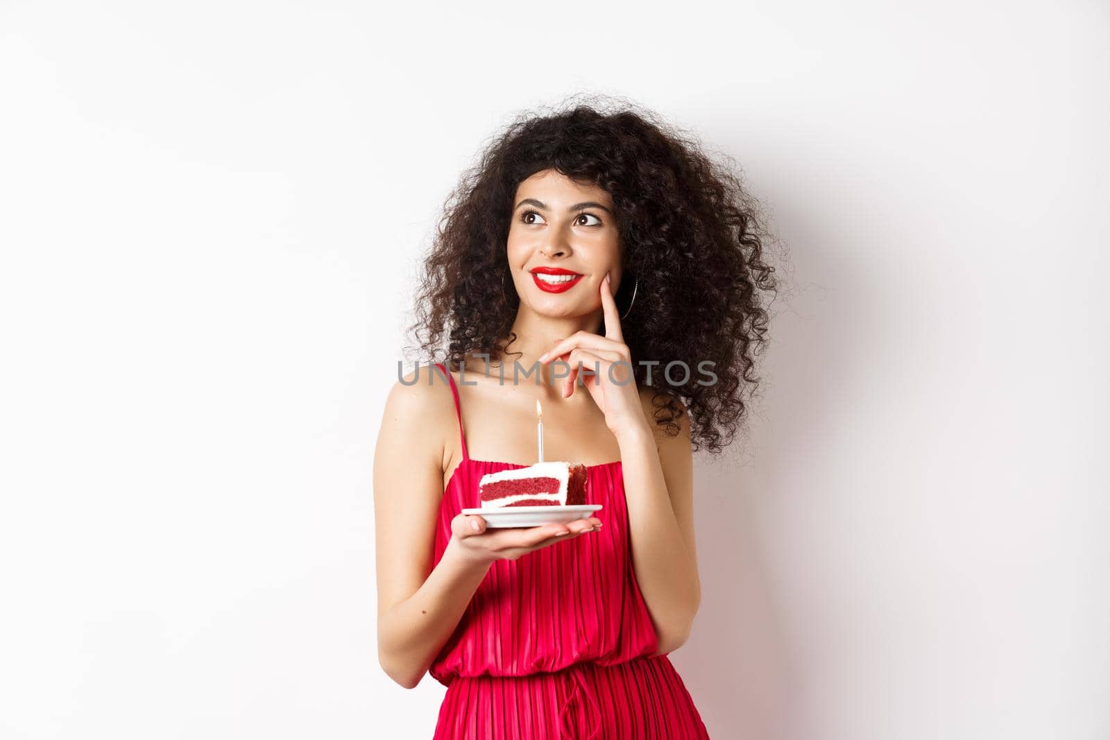 Happy woman celebrating birthday, holding cake and making wish, looking aside dreamy, standing in red dress on white background. Celebration and holiday concept by Benzoix