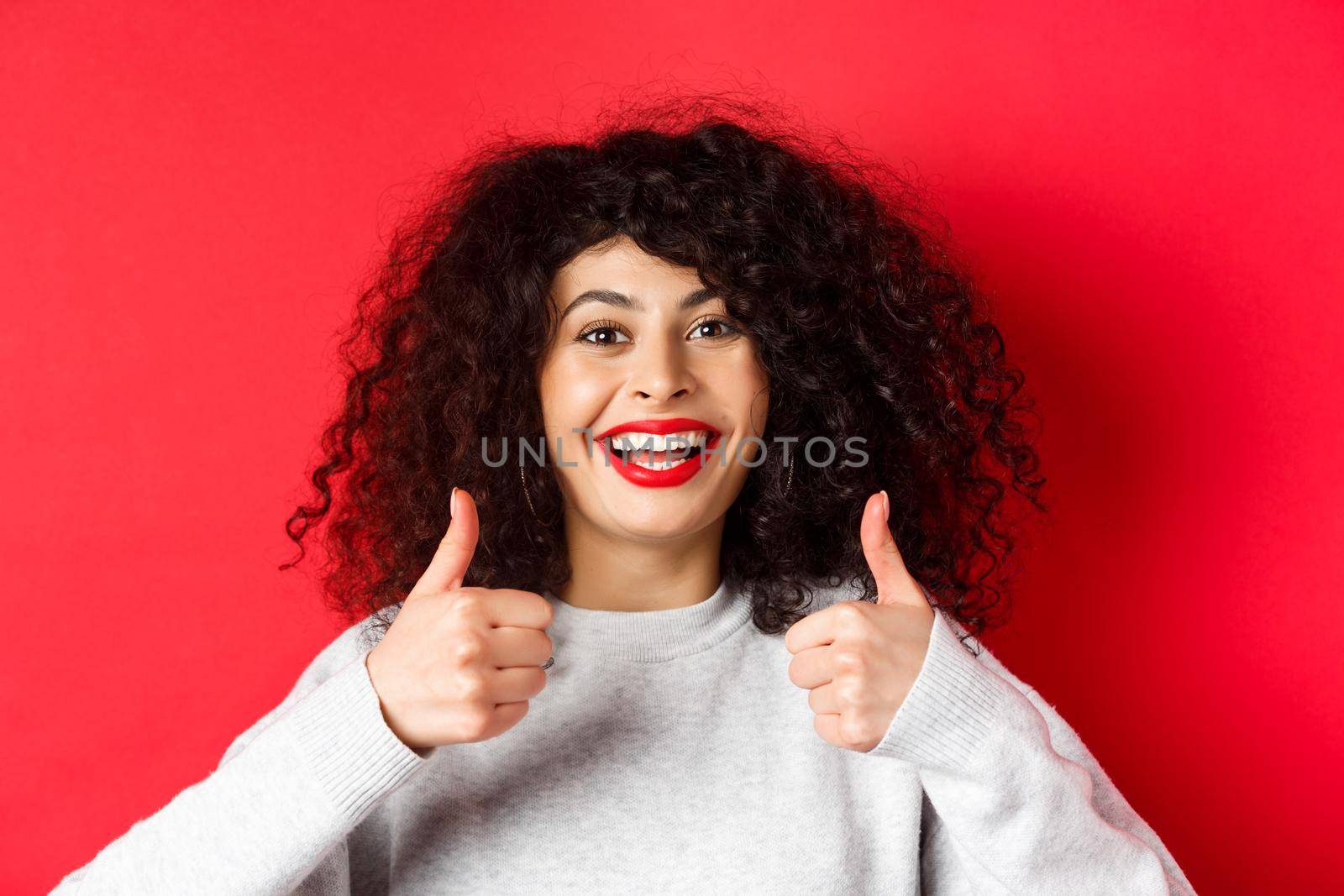 Close up of cheerful caucasian woman showing thumbs up in approval, smiling pleased, like and praise something good, standing on red background.