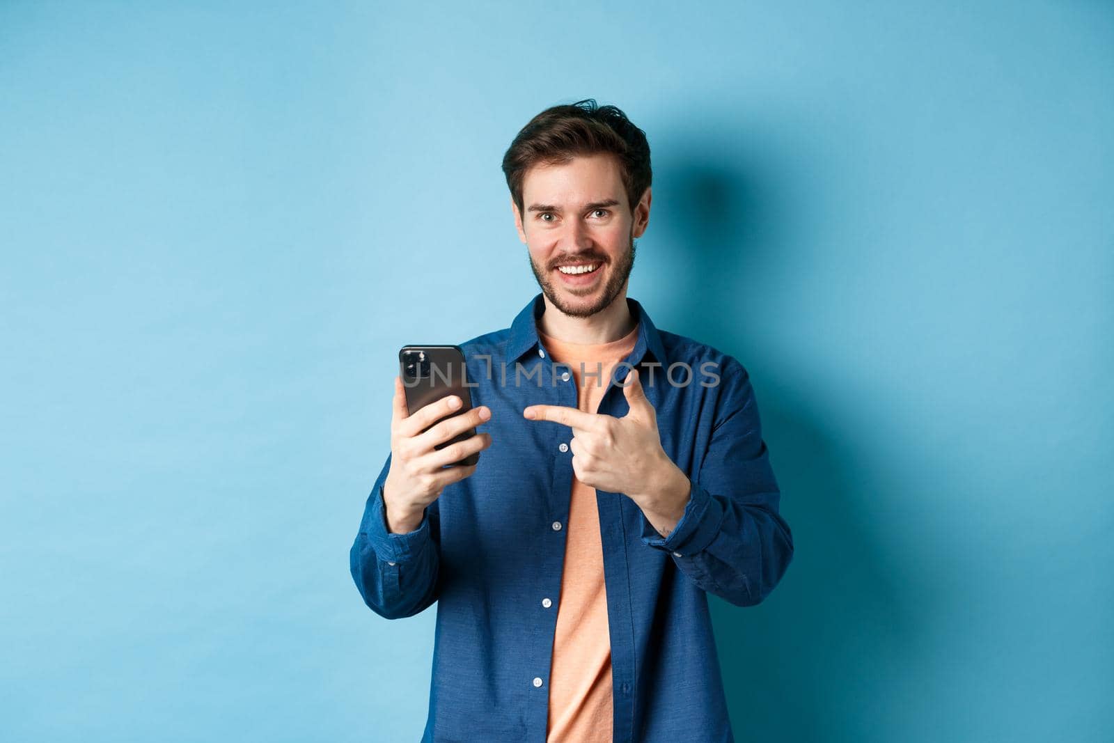 Handsome smiling man pointing finger at smartphone and looking pleased at camera, recommending app, standing on blue background by Benzoix