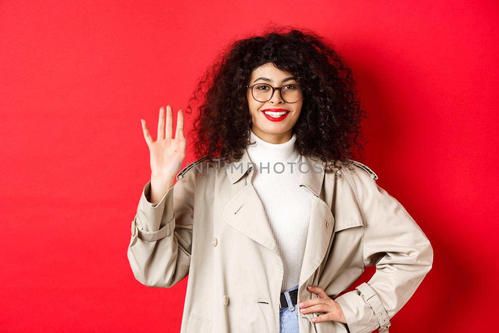 Stylish european woman in glasses and trench coat, waiving hand and smiling, saying hello, greeting someone, standing on red background.