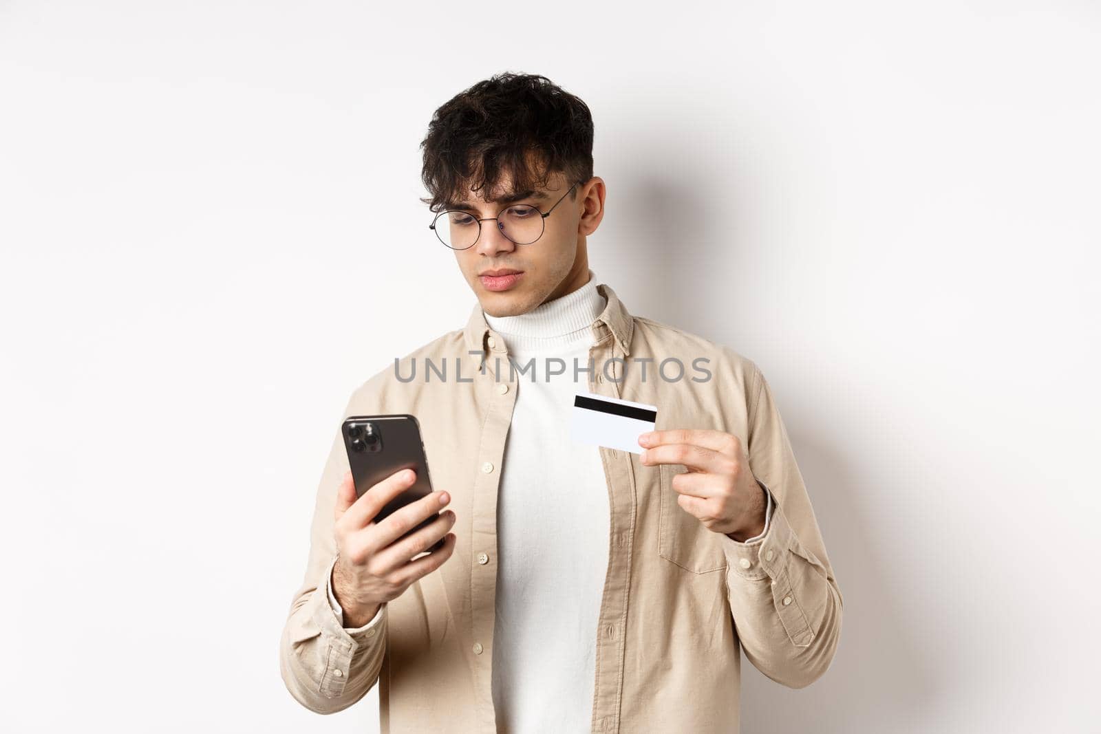 Handsome young man in glasses making purchase on phone, shopping online, holding plastic credit card and smartphone, standing on white background.