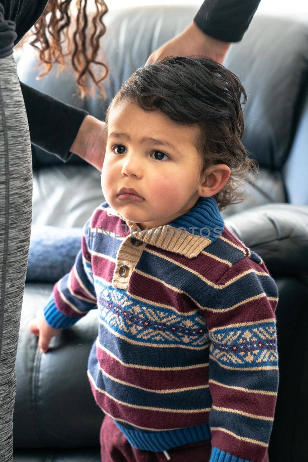 A handsome young baby boy or toddler with long curly brown hair wearing a patterned and striped collared sweater stands by his mother's leg as she combs and fixes the child's hair.