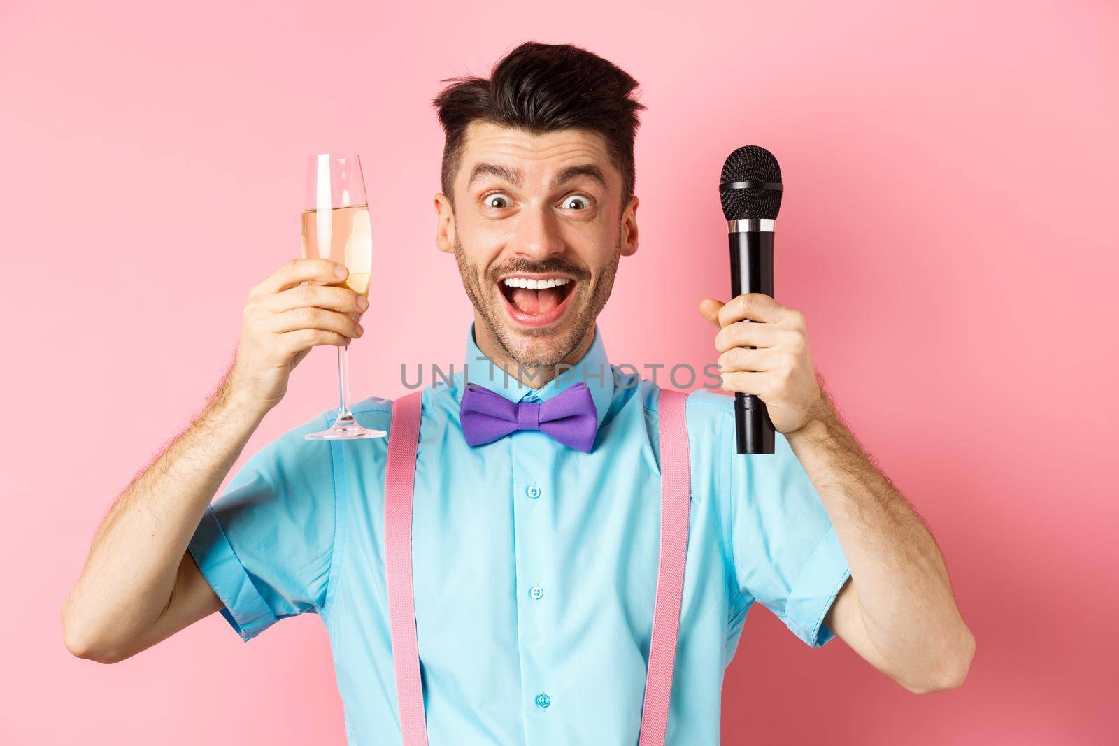 Party and festive events concept. Cheerful young male entertainer, giving speech on holiday, raising glass of chamapgne and holding microphone, making toast on wedding, pink background.