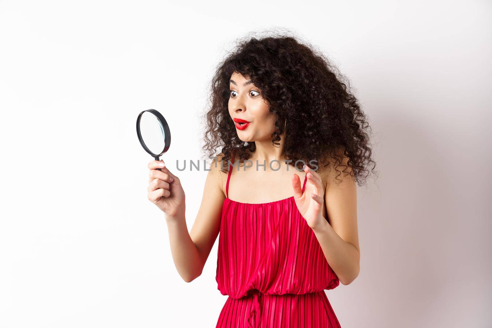 Excited woman in red dress look through magnifying glass and smiling, found interesting promo, standing on white background.