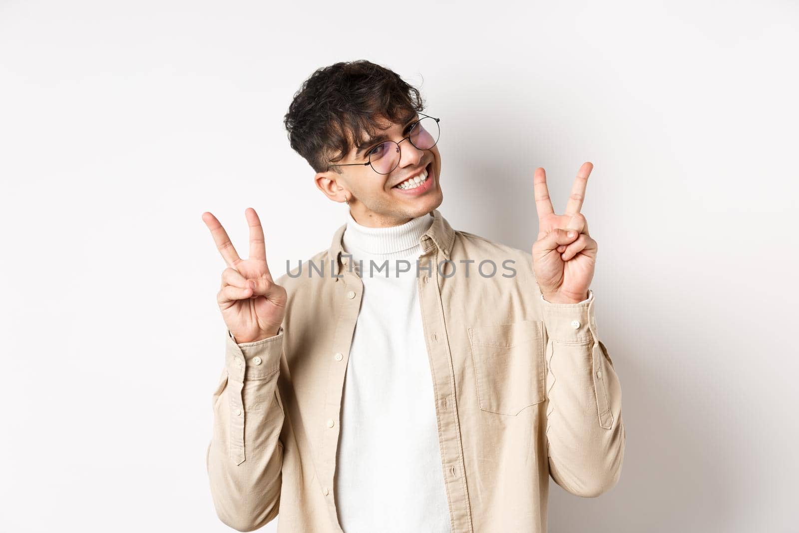 Real people. Handsome hipster guy in glasses showing peace gestures and smiling cute, standing on white background by Benzoix