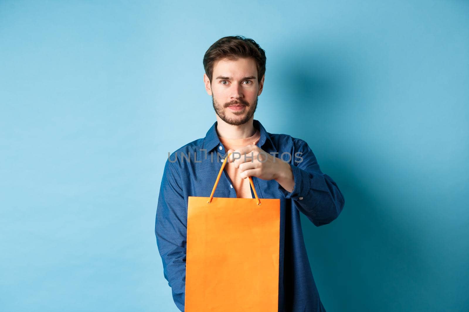 Handsome bearded man stretch out hand with orange shopping bag, making a gift, standing on blue background by Benzoix