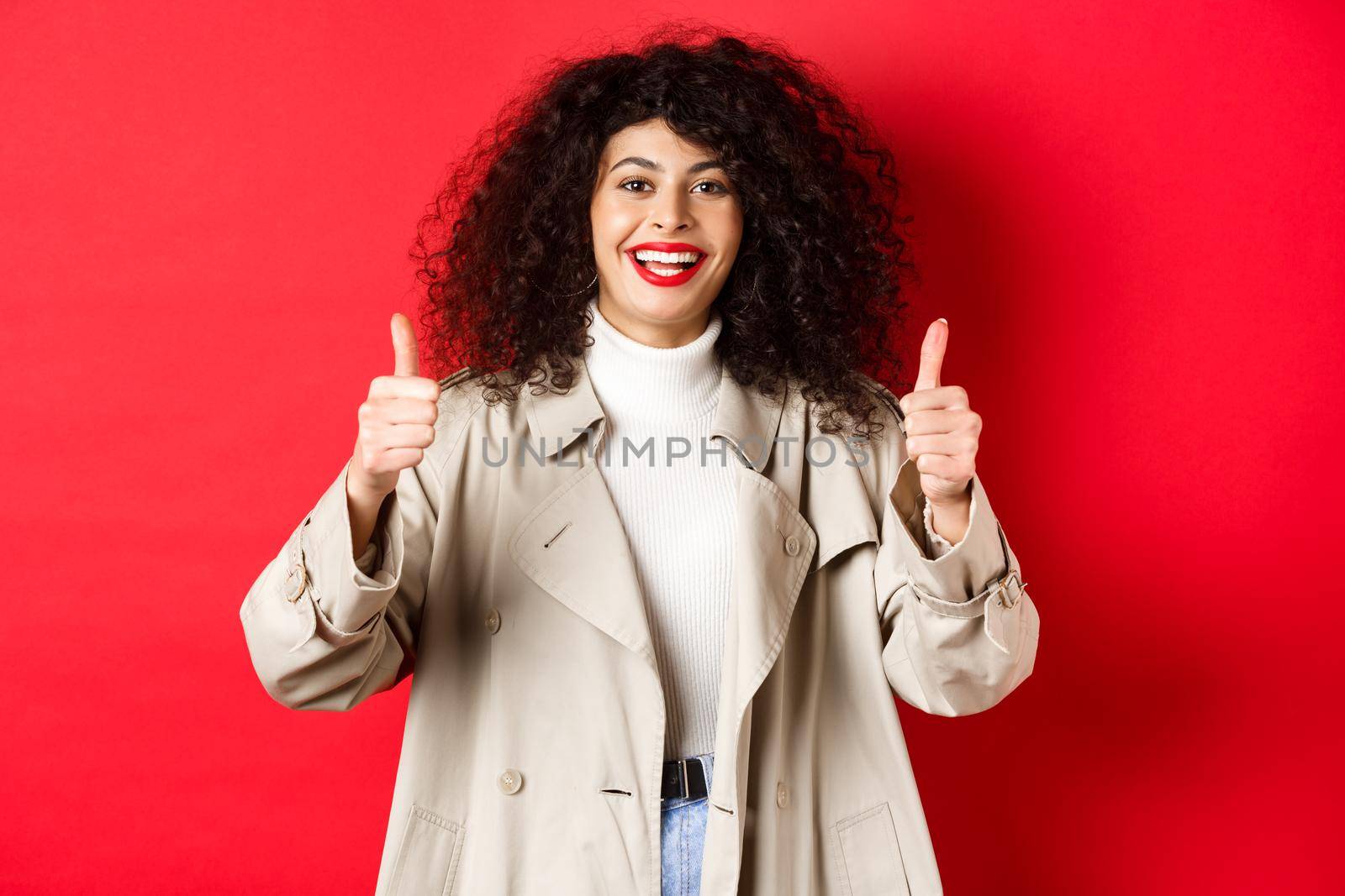 Portrait of young woman looking happy, wearing trench coat and showing thumbs-up, say yes, approve and praise something good, red background.