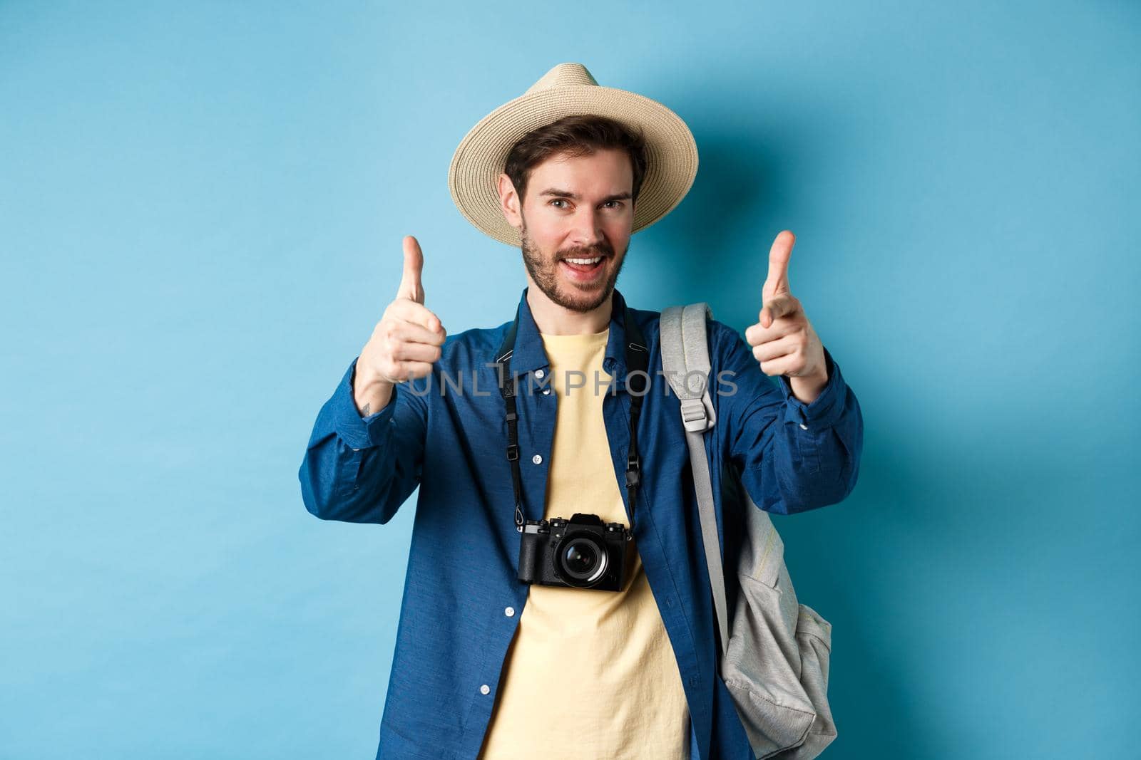 Handsome tourist going on summer vacation with camera and backpack pointing fingers at camera and inviting you, standing on blue background by Benzoix