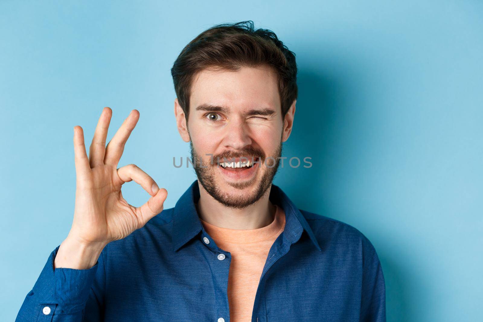 Close up of cheerful and confident bearded man assure everything good, showing okay gesture and winking, smiling at camera, standing on blue background.