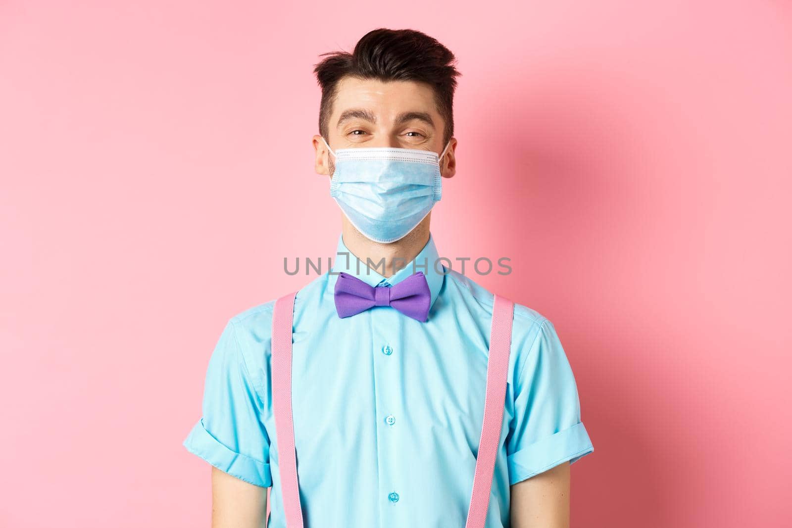 Covid-19, pandemic and health concept. Portrait of smiling man in medical mask feeling happy, standing in festive bow-tie and suspenders, pink background by Benzoix