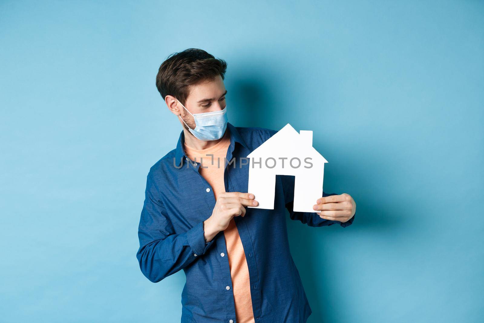 Real estate and quarantine concept. Young caucasian man in medical mask looking curious at paper house cutout, standing on blue background by Benzoix